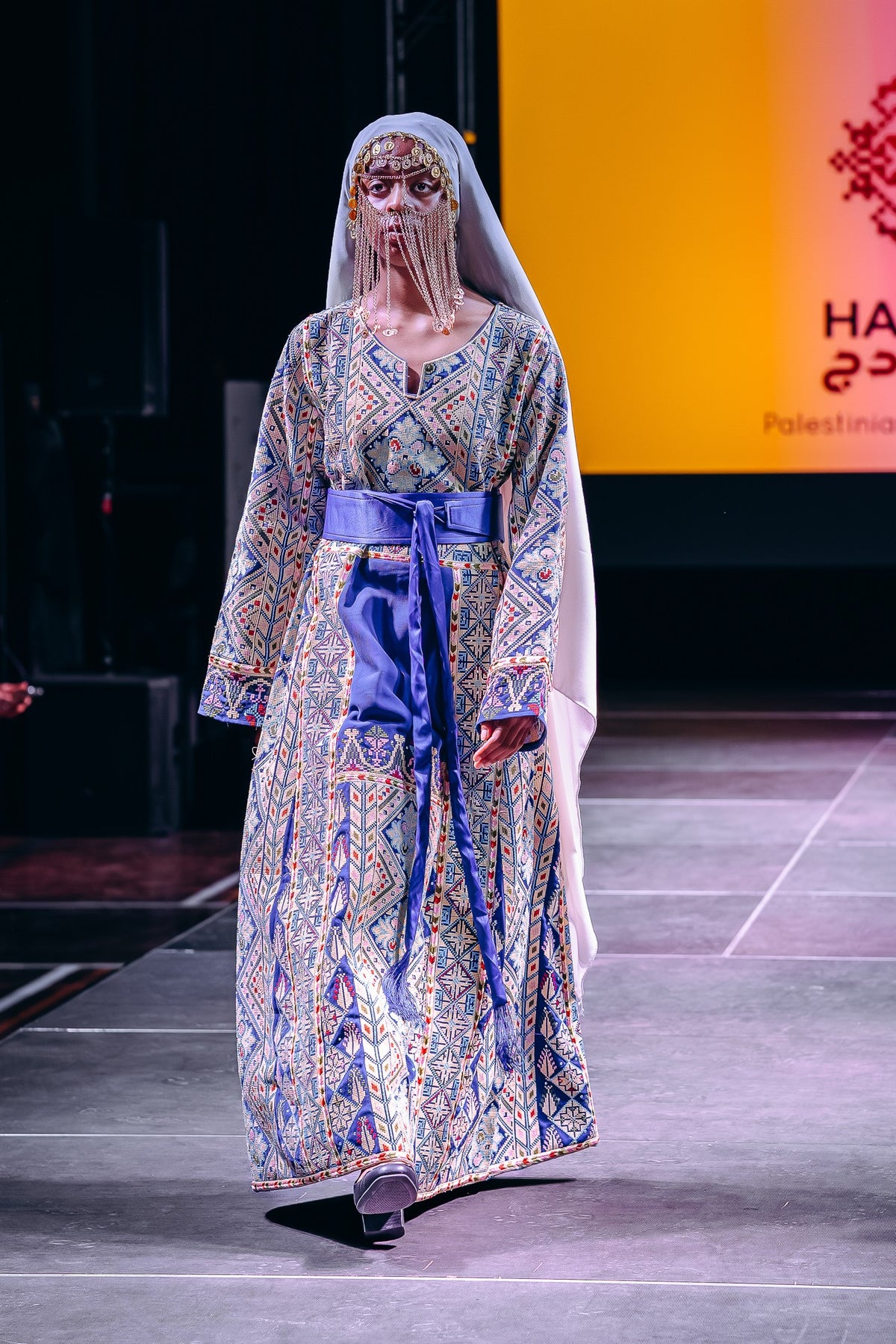A model walks the runway wearing a long, patterned dress with blue, gold, and beige tones, a wide blue waist sash, and a white veil. The background features a runway setting with a large orange screen displaying text.