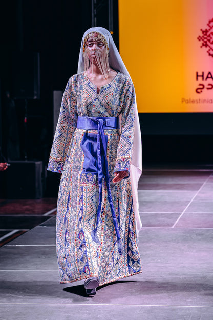 A model walks the runway wearing a long, patterned dress with blue, gold, and beige tones, a wide blue waist sash, and a white veil. The background features a runway setting with a large orange screen displaying text.