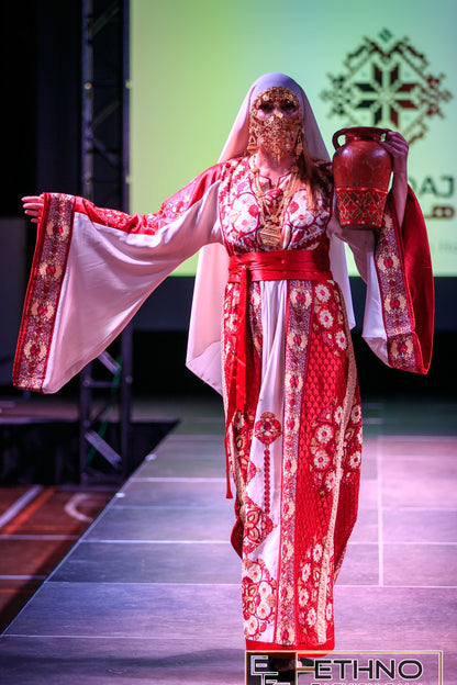 A model walks a fashion runway wearing a traditional red and white patterned robe with a matching head covering and face mask, holding a red decorative jug.