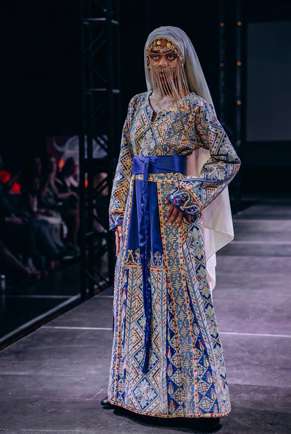 A model walks the runway wearing a long, ornate traditional dress with intricate blue, gold, and beige geometric patterns, a solid royal blue sash at the waist, and a sheer white veil. The photo is taken on a fashion gala runway with a dark stage background and audience blurred in the shadows.