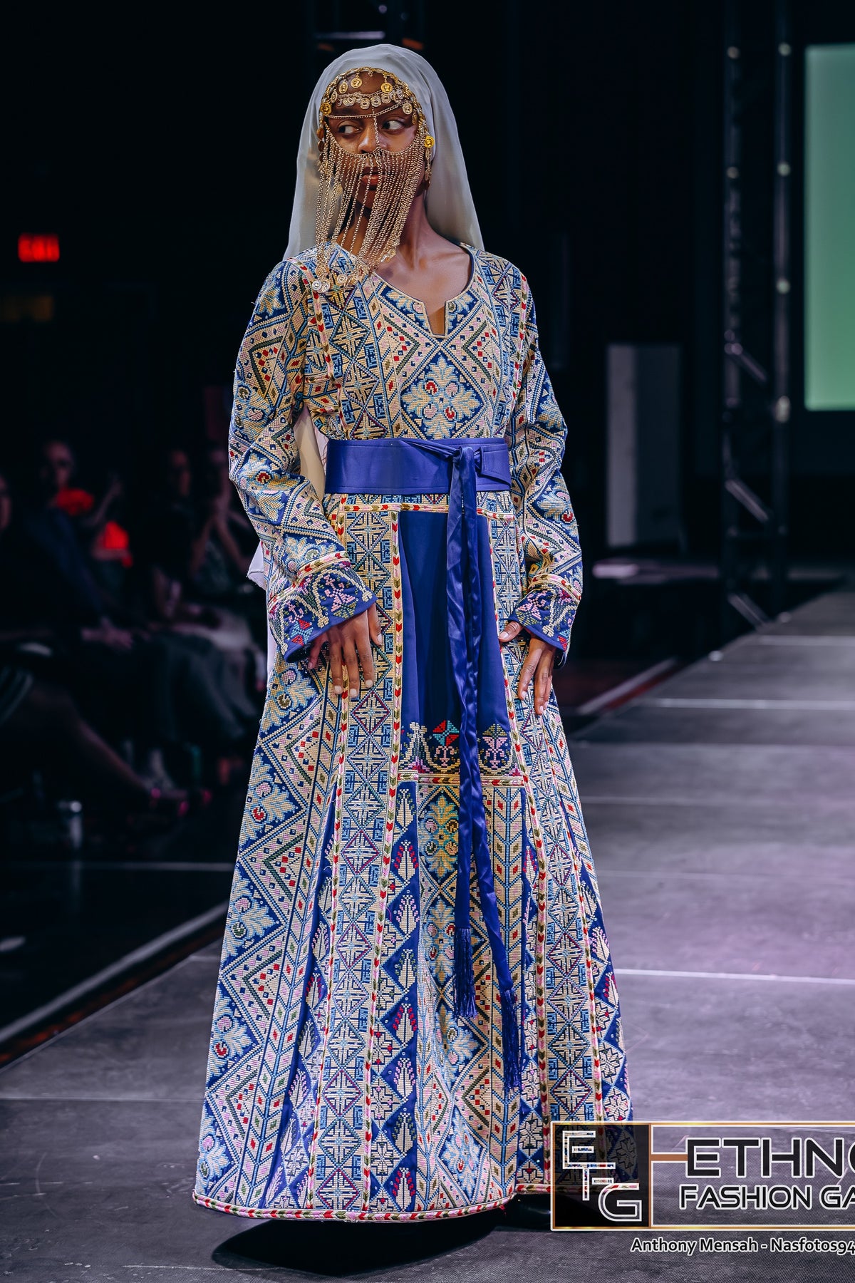 A model walks the runway wearing a long-sleeved, floor-length traditional dress with an intricate blue, yellow, and white geometric pattern, a solid royal blue sash at the waist, and a sheer white veil with gold face detailing.