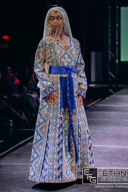 A model walks the runway wearing a long-sleeved, floor-length traditional dress with an intricate blue, yellow, and white geometric pattern, a solid royal blue sash at the waist, and a sheer white veil with gold face detailing.